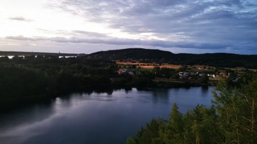 Turkusowe Jezioro otoczone lasami, z panoramą wsi w oddali i zachodzącym słońcem.